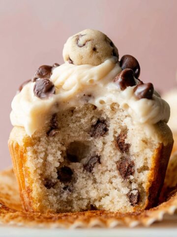 Delicious Molten Cookie Dough Cupcakes 8 A freshly baked Molten Cookie Dough Cupcakes, topped with smooth buttercream frosting, mini chocolate chips, and a ball of cookie dough. The cupcake is partially unwrapped to reveal the soft interior filled with chocolate chips.