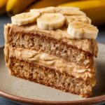 A slice of moist Peanut Butter Banana Cake with creamy peanut butter frosting, topped with fresh banana slices, showcasing the soft texture and layered appearance of the cake.