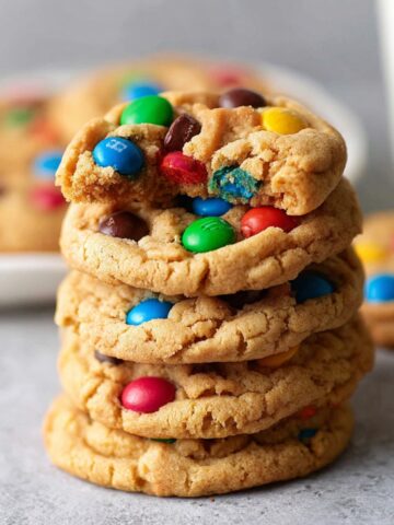 Soft Peanut Butter M&M Cookies Recipe 2 Freshly baked Peanut Butter M&M Cookies stacked high on a plate, with one cookie broken to reveal its soft, gooey center.