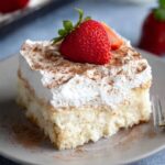 A slice of Tres Leches Cake topped with whipped cream, a sprinkle of cinnamon, and a fresh strawberry garnish.