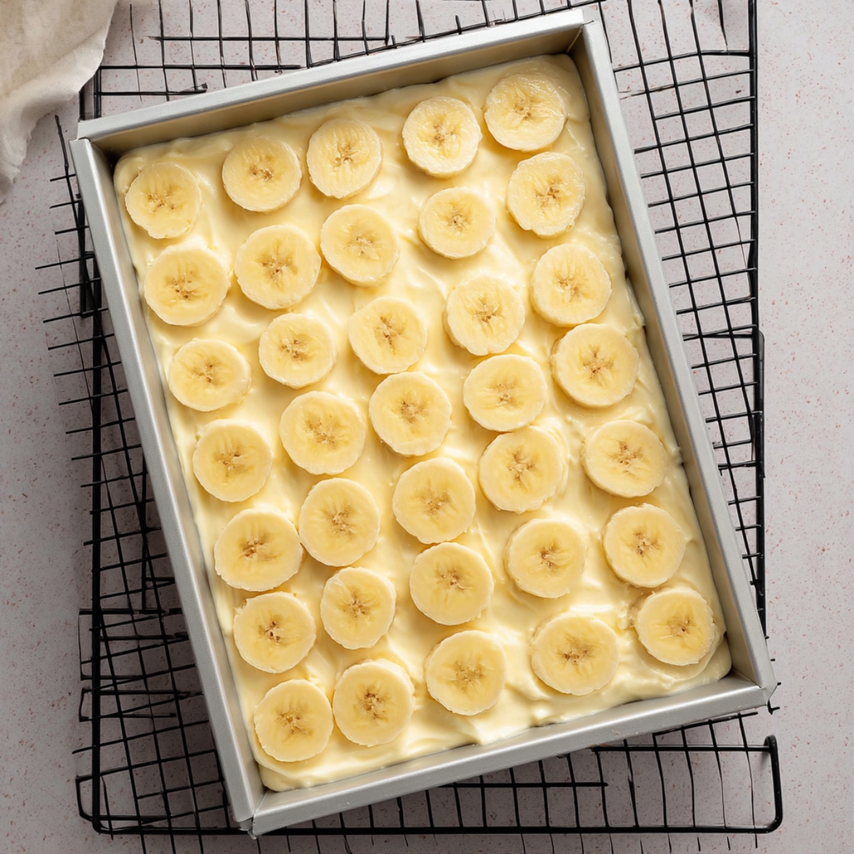 A baking dish with a layer of banana slices on top of a creamy banana pudding mixture. The dish is ready for the next step in making banana cream bars.