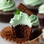 A freshly baked Andes Mint Cupcakes topped with mint-green frosting and a piece of Andes mint chocolate. A bite has been taken out, revealing the moist chocolate interior.