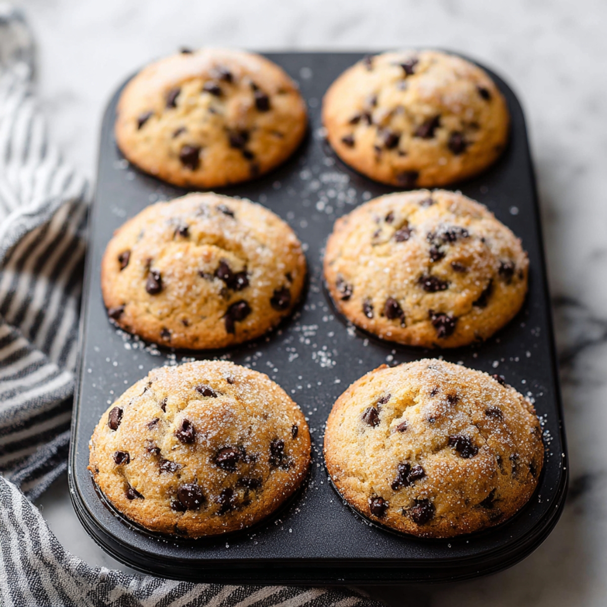 Easy Chocolate Chip Muffins Recipe 11 Six freshly baked chocolate chip muffins with a light sprinkle of sugar, placed in a muffin pan, with visible chocolate chips on top.