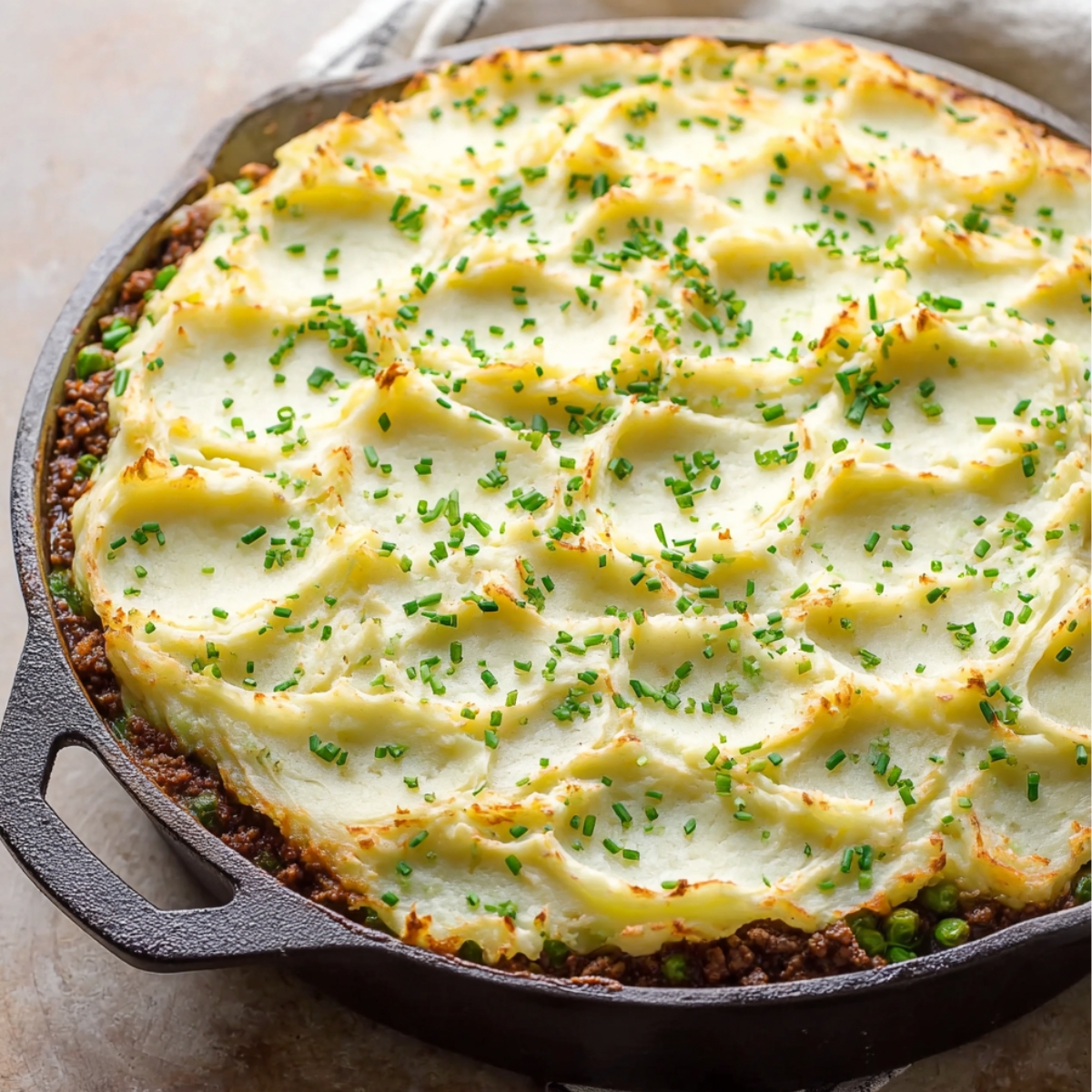 Cottage Pie with Garlic Mashed Potatoes 11 A golden and crispy layer of mashed potatoes atop a savory cottage pie with ground beef, peas, and carrots, garnished with fresh chives in a rustic cast iron skillet.
