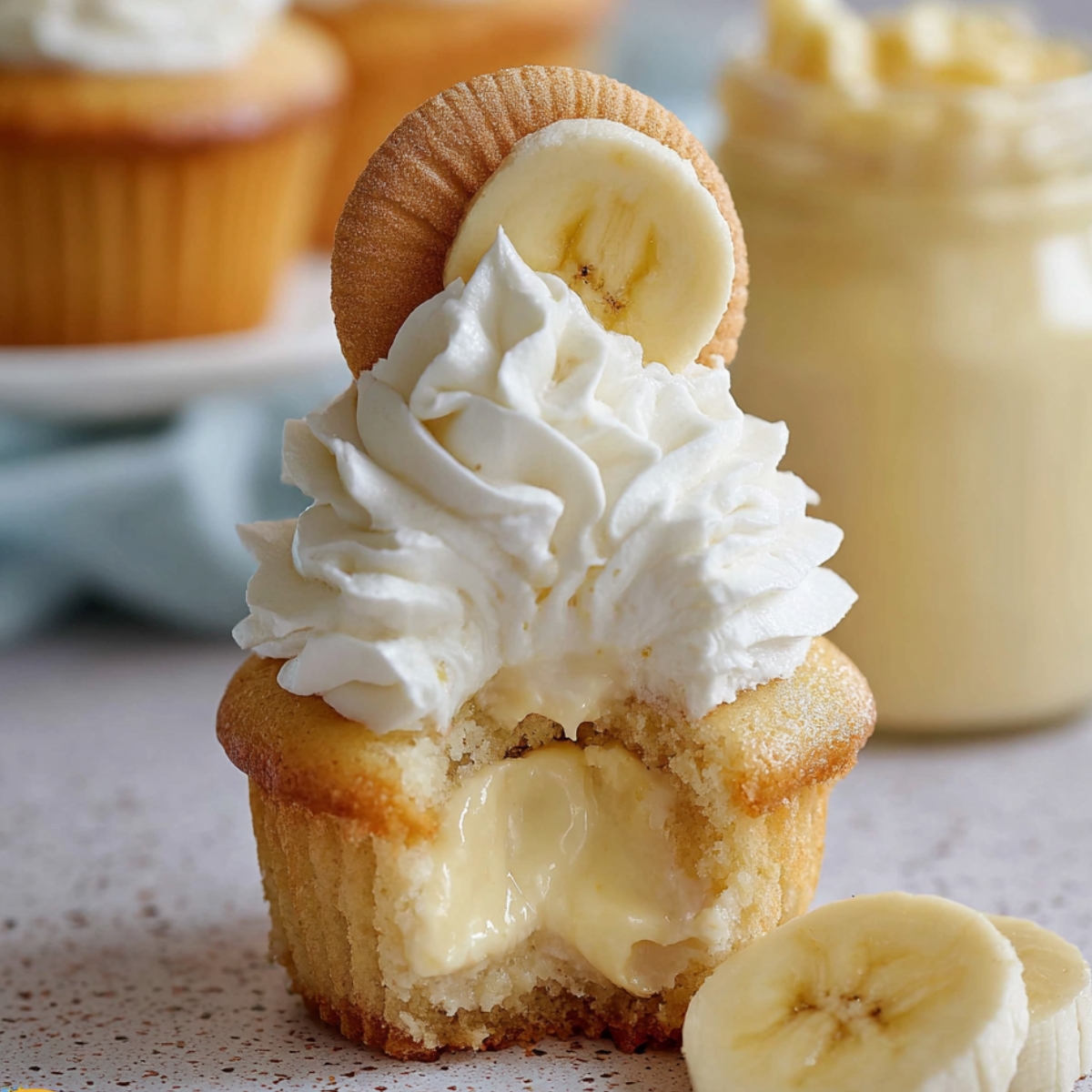 A Banana Pudding Cupcakes topped with whipped cream, a vanilla wafer, and a slice of banana, with the cupcake cut open to reveal a creamy banana filling inside.