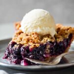 A slice of Blueberry Crumble Pie served on a plate with a scoop of vanilla ice cream on top, showing the vibrant blueberry filling and golden crumble topping.