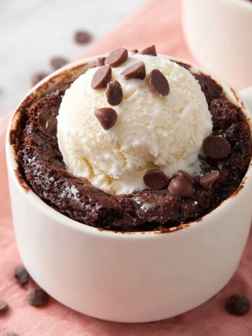 Easy Brownie in a Mug Recipe 2 A mug filled with a freshly baked Brownie in a Mug topped with vanilla ice cream and chocolate chips, placed on a pink napkin.