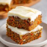 Two stacked carrot cake bars with cream cheese frosting, showing the moist texture with grated carrots and nuts inside, served on a plate.