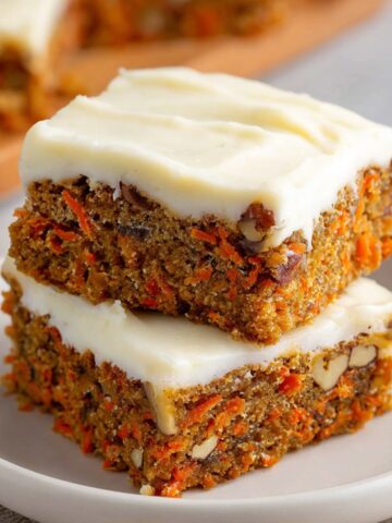 Two stacked carrot cake bars with cream cheese frosting, showing the moist texture with grated carrots and nuts inside, served on a plate.