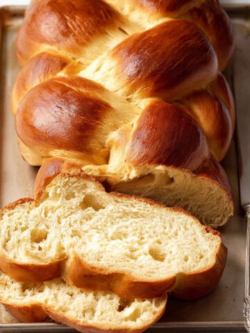 The Best Challah Bread Recipe 1 Freshly baked, golden-brown braided challah bread on a baking tray, with a slice cut to reveal the fluffy, airy interior.