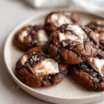 A plate of soft Chocolate Marshmallow Swirl Cookies topped with melted marshmallow swirls, dark chocolate, and a sprinkle of sea salt.