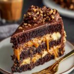 A decadent slice of Chocolate Snickers Cake, with layers of rich chocolate cake, creamy caramel, and smooth peanut butter frosting, topped with crushed peanuts.