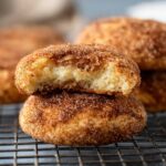 A close-up of a freshly baked Churro Cheesecake Cookies, split in half to reveal its soft, creamy interior. The cookie is coated with cinnamon sugar on the outside.