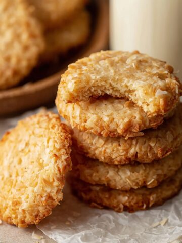 The Best Coconut Biscuits Recipe 1 Freshly baked Coconut Biscuits stacked together, golden and crispy on the edges with a soft, chewy interior. One biscuit has a bite taken out, revealing its texture.