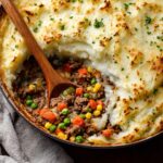 A warm, comforting Cottage Pie with Garlic Mashed Potatoes on top, revealing a hearty filling of seasoned ground beef and vegetables like peas, corn, and carrots, with a wooden spoon serving a portion.