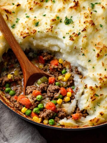 A warm, comforting Cottage Pie with Garlic Mashed Potatoes on top, revealing a hearty filling of seasoned ground beef and vegetables like peas, corn, and carrots, with a wooden spoon serving a portion.
