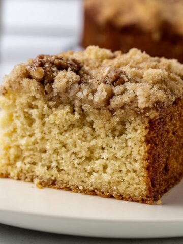A close-up of a moist, crumbly slice of Cowboy Coffee Cake with a crumbly topping, sitting on a white plate. The cake is golden in color with a slightly crunchy topping, indicating a delicious, homemade treat.