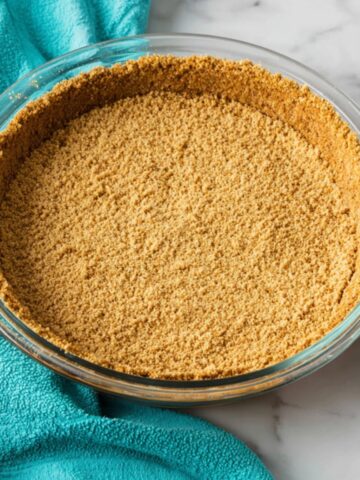 Easy Graham Cracker Crust Recipe 2 A glass pie dish filled with a Graham Cracker Crust, neatly pressed and evenly distributed around the base and sides. The crust is surrounded by a teal kitchen towel with graham crackers placed nearby.