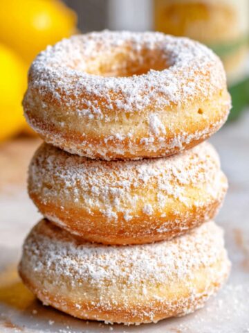 Greek Yogurt Cake Donuts Recipe 5 A stack of three freshly baked, golden Greek Yogurt Cake Donuts dusted with powdered sugar, set against a soft, lemony background.