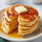 A stack of fluffy, golden-brown Greek Yogurt Pancakes topped with a square of melting butter, served on a plate. The pancakes are stacked high, with a rich golden-brown color and a soft interior.