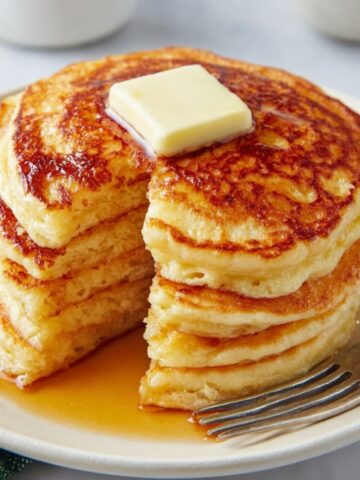 A stack of fluffy, golden-brown Greek Yogurt Pancakes topped with a square of melting butter, served on a plate. The pancakes are stacked high, with a rich golden-brown color and a soft interior.