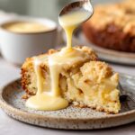 Close-up image of a slice of Irish apple cake with creamy custard being poured over the top. The cake has a golden crust and soft, fluffy interior, served on a speckled plate.
