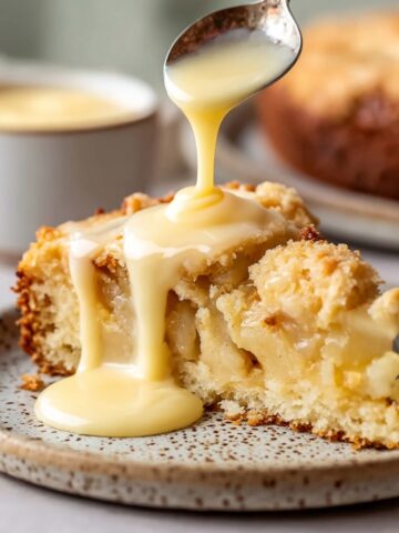 Traditional Irish Apple Cake Recipe 1 Close-up image of a slice of Irish apple cake with creamy custard being poured over the top. The cake has a golden crust and soft, fluffy interior, served on a speckled plate.