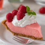 A close-up shot of a slice of pink Jello pie topped with whipped cream and garnished with fresh raspberries.
