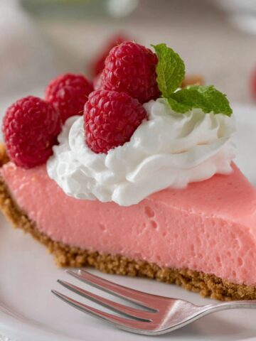A close-up shot of a slice of pink Jello pie topped with whipped cream and garnished with fresh raspberries.