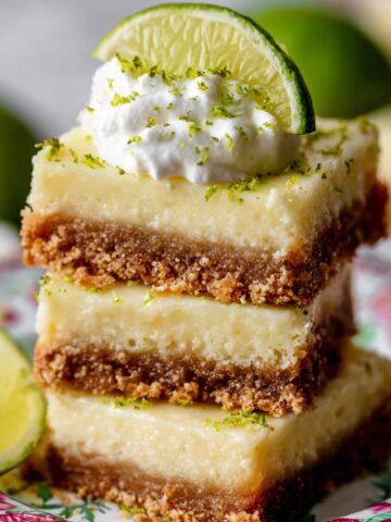 A stack of Key Lime Pie Bars with a graham cracker crust, topped with whipped cream, lime zest, and a lime wedge, served on a floral plate.