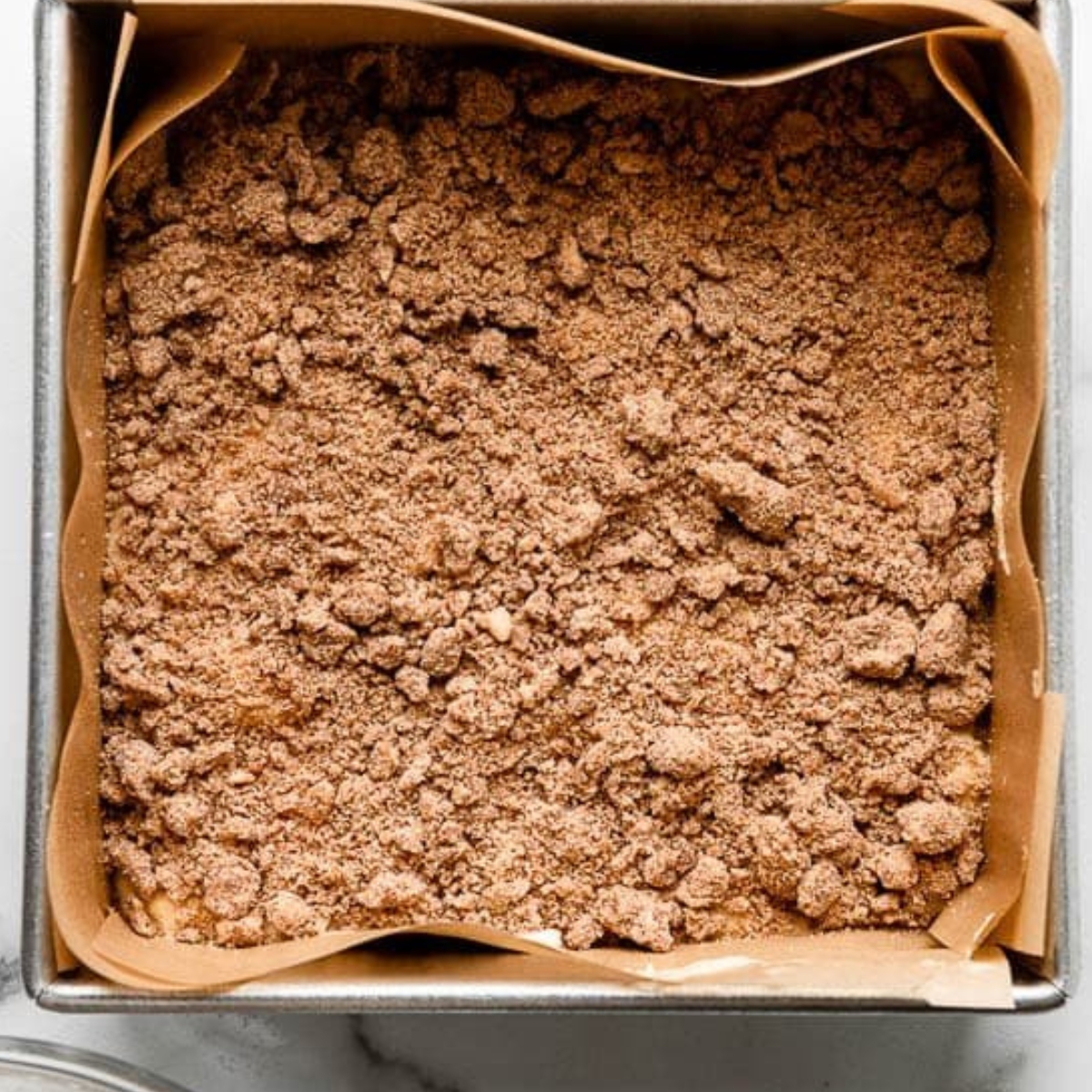 Sour Cream Coffee Cake Recipe 11 A square baking pan lined with parchment paper, filled with a crumbled cinnamon streusel topping, ready to be baked.