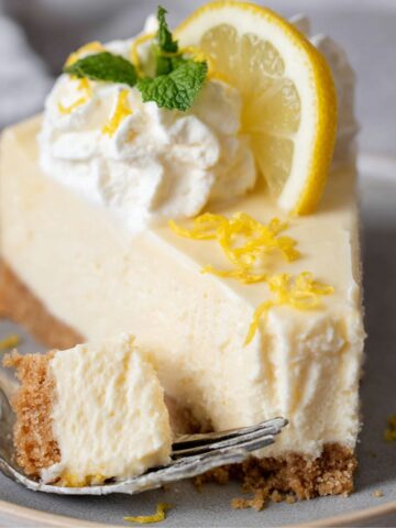 A close-up of a slice of Lemon Greek Yogurt Cheesecake topped with whipped cream, a lemon slice, and lemon zest, served with a forkful of the creamy dessert.