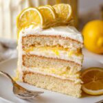 A slice of lemon layer cake on a white plate showing fluffy layers of cake, lemon curd filling, and cream cheese frosting, garnished with fresh lemon slices on top.
