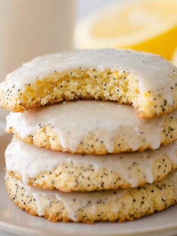 A close-up of stacked, glazed Lemon Poppy Seed Cookies with a bite taken out of the top cookie, showcasing the soft, crumbly texture.