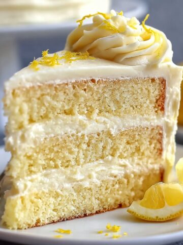 A slice of Lemon Velvet Cake with layers of creamy lemon frosting, garnished with lemon zest and served with lemon wedges on a white plate.