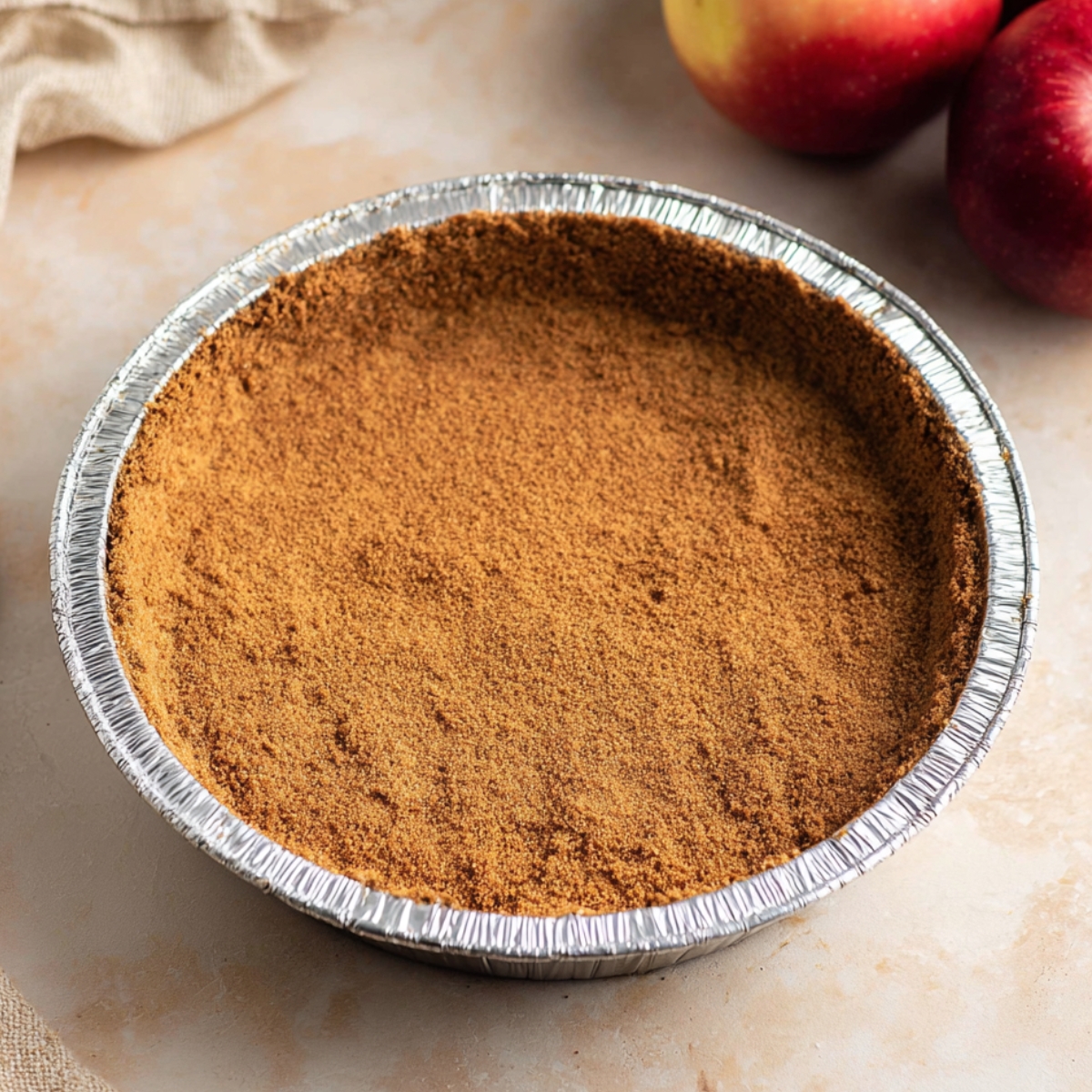 A gingersnap crust pressed into a pie pan, ready for filling, with fresh apples placed nearby.