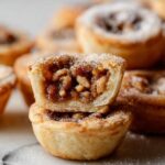 A stack of Maple Walnut Tassies, with one cut open to show the nutty filling. The crumbly pastry is lightly dusted with powdered sugar.
