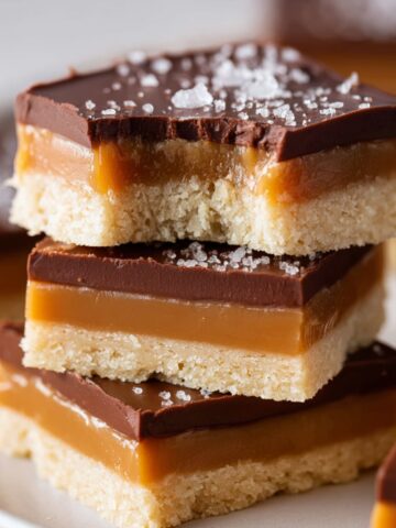Stack of millionaire shortbread bars with layers of shortbread, caramel, and chocolate, topped with sea salt on a plate.