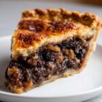 A close-up of a slice of Mincemeat Pie on a plate, showing the rich, dark filling of dried fruits and spices, with a golden-brown, flaky crust.