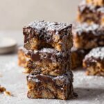 Three stacked muddy buddy bars, dusted with powdered sugar, showcasing the chocolate and cereal layers with a soft, crumbly texture.