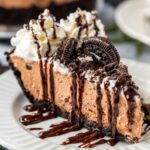 A decadent slice of mudslide pie on a white plate, topped with whipped cream, Oreo cookie pieces, and a drizzle of chocolate sauce, showcasing a rich and creamy texture.