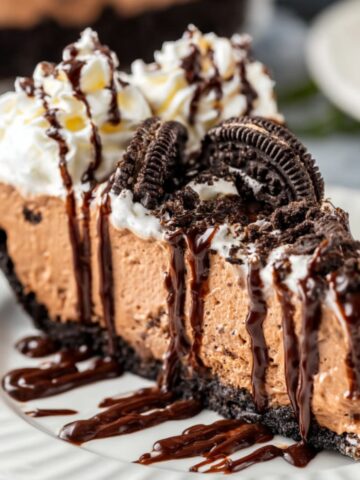 A decadent slice of mudslide pie on a white plate, topped with whipped cream, Oreo cookie pieces, and a drizzle of chocolate sauce, showcasing a rich and creamy texture.
