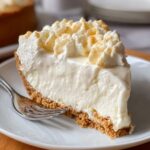 A slice of No Bake Marshmallow Cheesecake with a graham cracker crust, topped with whipped cream and a smooth creamy filling, served on a plate.