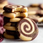 A stack of freshly baked chocolate-vanilla Pinwheel Cookies with a bite taken out of one, revealing the swirl inside. The cookies are dipped in dark chocolate and sprinkled with gold dust.