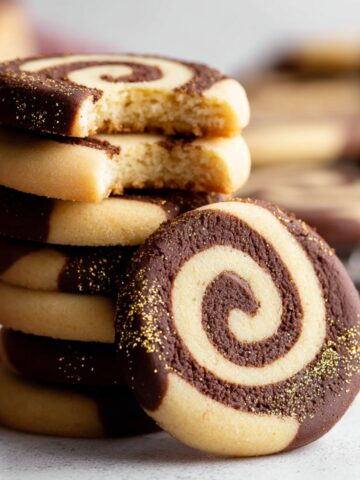 The Best Pinwheel Cookies Recipe 3 A stack of freshly baked chocolate-vanilla Pinwheel Cookies with a bite taken out of one, revealing the swirl inside. The cookies are dipped in dark chocolate and sprinkled with gold dust.