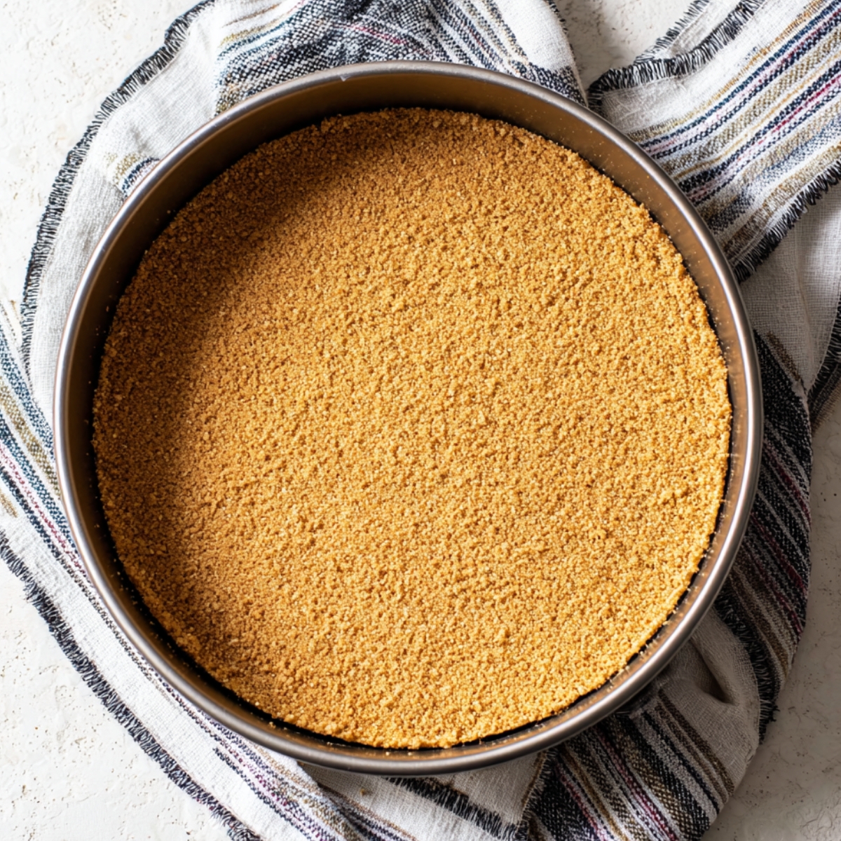 A graham cracker crust pressed into the bottom of a round springform pan, evenly spread and ready for the filling.
