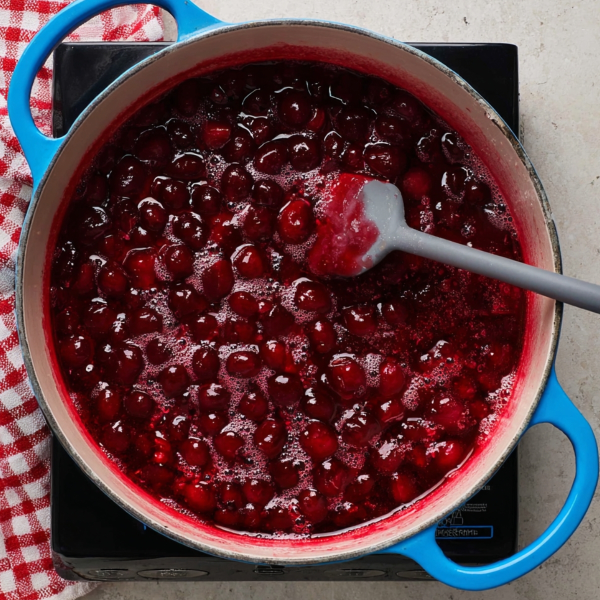 A saucepan with sour cherry pie filling simmering, with bright red cherries and syrup being stirred with a spatula.