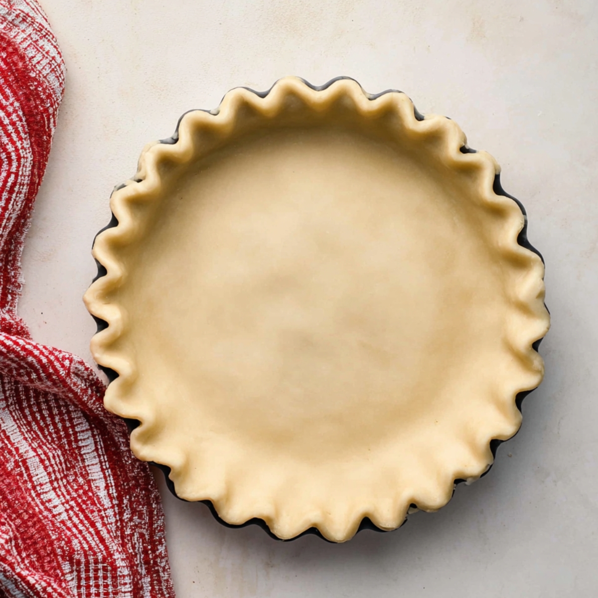 An unbaked pie crust with a crimped edge, resting in a pie dish, next to a red-and-white checkered towel.