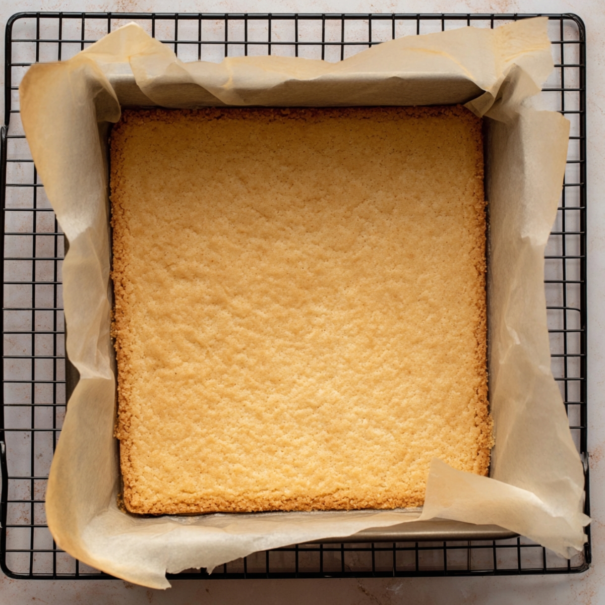Freshly baked shortbread crust in a square baking pan, cooling on a wire rack, lined with parchment paper.