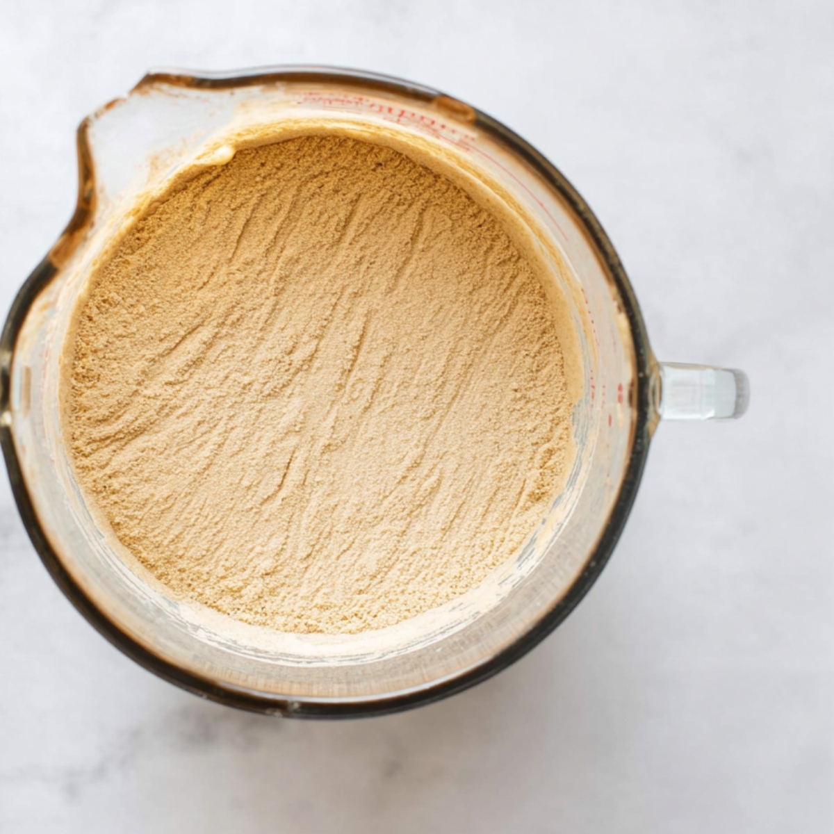 The Best Challah Bread Recipe 10 Clear glass measuring cup filled with dry yeast powder spread evenly at the bottom, with a clean white marble background.
